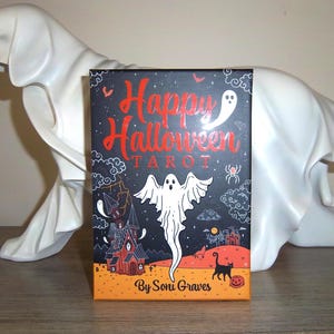 Puede incluir: Una baraja de cartas del tarot con temática de Halloween con un diseño negro y naranja. La caja presenta las palabras "Happy Halloween Tarot" en rojo, junto con ilustraciones de fantasmas, una casa embrujada y un gato negro. La baraja es de Soni Graves.