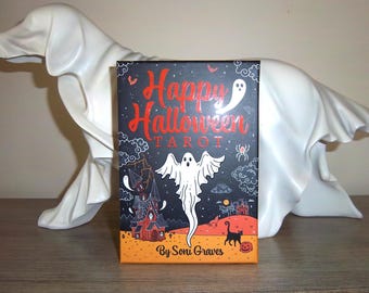 May include: A Halloween-themed tarot card deck with a black and orange design. The box features the words "Happy Halloween Tarot" in red, along with illustrations of ghosts, a haunted house, and a black cat. The deck is by Soni Graves.