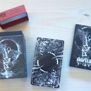 May include: An overhead shot of a deck of cards and its box. The box and cards are black with white graphic designs. The text "qnelia" is visible on the cards and a red box. The cards and boxes are on a light wood surface.