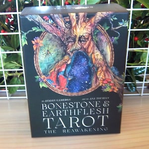 May include: A tarot card box with a detailed illustration of a tree-like face within a circular frame. The box is black with the words "BONESTONE & EARTHFLESH TAROT" and "THE REAWAKENING" in silver lettering. The artwork features vibrant colors.