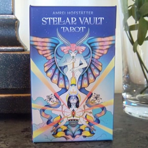 Może przedstawiać: Pudełko kart tarota "STELLAR VAULT TAROT" autorstwa AMREI HOFSTÄTTER na niebieskim tle. Pudełko zawiera ilustrację niebiańskiej postaci ze skrzydłami motyla i kobietą w pozycji medytacyjnej, w żywych kolorach.
