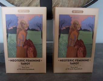 Può includere: Due scatole del mazzo di Tarot Femminile Neoterico. Le scatole sono beige con un bordo marrone chiaro. La parte anteriore di ogni scatola presenta una donna con i capelli biondi in piedi in un campo con un animale marrone. Il testo sulla scatola recita "WYSPELL +NEOTERIC FEMININE+ TAROT Mazzo di tarot di 78 carte e guida".
