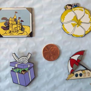 Puede incluir: Cuatro pines de esmalte con diseños de playa. Uno representa un castillo de arena en una playa amarilla, otro una gran flor blanca y amarilla con personajes, una caja de regalo morada y una sombrilla de playa. Se incluye una moneda.