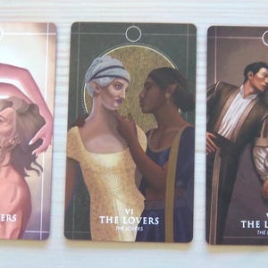May include: Three tarot cards depicting "The Lovers" card. Each card features a different illustration of two people embracing. The first card shows a woman with long dark hair embracing a man with short brown hair. The second card shows a woman with short blonde hair embracing a woman with long dark hair. The third card shows two men embracing. Each card has the Roman numeral "VI" and the words "The Lovers" printed on it.