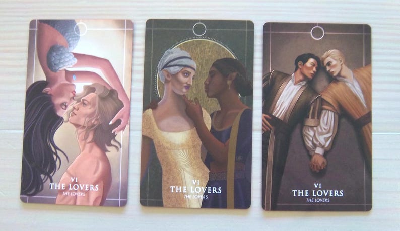 May include: Three tarot cards depicting "The Lovers" card. Each card features a different illustration of two people embracing. The first card shows a woman with long dark hair embracing a man with short brown hair. The second card shows a woman with short blonde hair embracing a woman with long dark hair. The third card shows two men embracing. Each card has the Roman numeral "VI" and the words "The Lovers" printed on it.