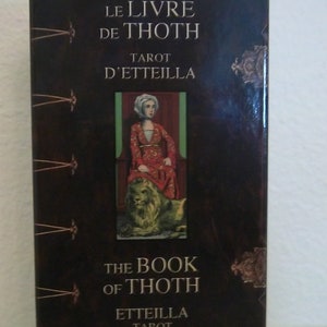 Op de afbeelding: Een bruine doos met gouden accenten heeft de tekst "Le Livre de Thoth Tarot d'Etteilla" en "The Book of Thoth Etteilla Tarot". De doos heeft een afbeelding van een vrouw in een rode jurk die op een leeuw zit.