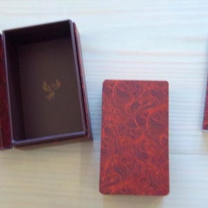 May include: A red box with a gold design and the text "Embark on a Journey Through the Eye of the Divine" and "A Selection of the Diviner Within All of Us". The box is open and shows a smaller box inside. A red deck of cards with a gold design is next to the box. A small booklet with a white background and the text "Cosmic Seeker Tarot" is on the right side of the image.