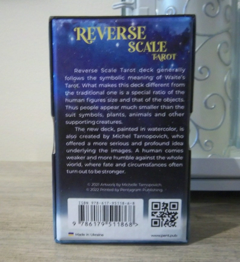 May include: A blue and gold box with the text "Reverse Scale Tarot" on the front. The back of the box has a description of the tarot deck, the ISBN number, and a QR code. The box is made in Ukraine.