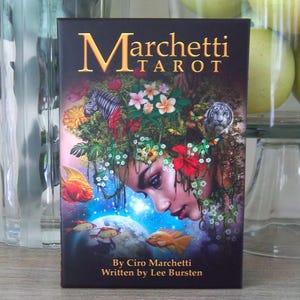 May include: A tarot card deck box with the title "Marchetti Tarot" in gold lettering. The artwork features a woman's face with floral and animal elements, including a zebra, tiger, and goldfish. The text "By Ciro Marchetti Written by Lee Bursten" is at the bottom.