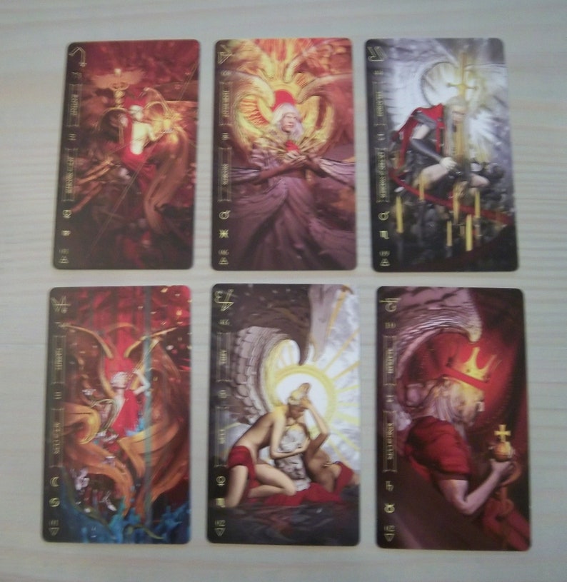 May include: Six tarot cards with gold accents and illustrations of angels. The cards are in a vertical row, with the top card showing a red and gold angel with wings spread wide. The second card shows a white angel with wings spread wide. The third card shows a black and white angel with wings spread wide. The fourth card shows a red and gold angel with wings spread wide. The fifth card shows a white angel with wings spread wide. The sixth card shows a black and white angel with wings spread wide.