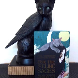 May include: A black winged cat figurine with green eyes stands behind a rectangular deck of cards titled Ask the Feline Sages, featuring an illustration of a person with a grey cat on their head against a dark teal background with a moon.