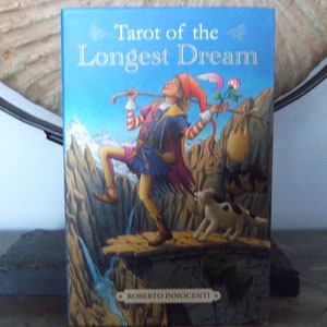 May include: A book cover with a blue background and the title "Tarot of the Longest Dream" in white lettering. The cover features an illustration of a jester-like figure walking on a narrow path with a dog and a bag.