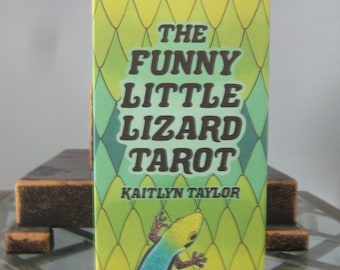 May include: A yellow and green book with a diamond pattern on the cover. The book is titled "The Funny Little Lizard Tarot" by Kaitlyn Taylor. A green and blue lizard is pictured on the bottom of the cover.