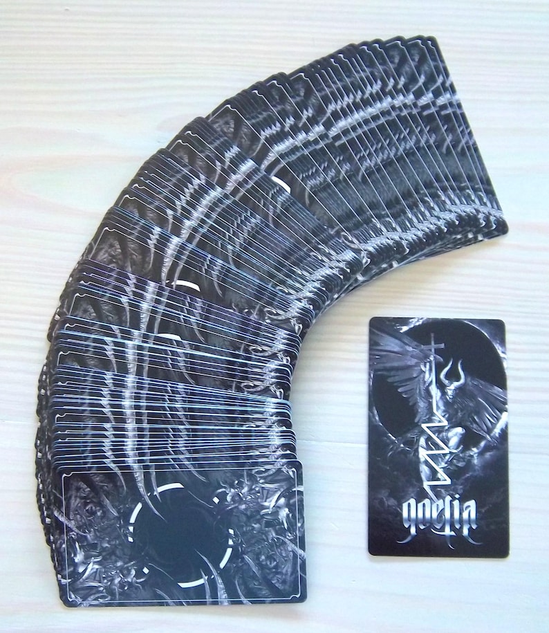 May include: A deck of tarot cards with a dark, gothic aesthetic. The cards are predominantly black and white, featuring intricate illustrations and the word "goetia" on one card. The cards are fanned out, with one card displayed separately.