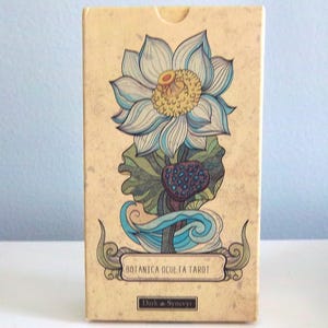 Puede incluir: Una caja de tarot con fondo beige e ilustración floral. La ilustración presenta una gran flor de loto azul claro y amarillo con hojas verdes y una vaina de semillas azul oscuro. Se muestra el texto "BOTANICA OCULTA TAROT".