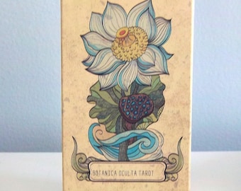 May include: A tarot card box with a beige background and a floral illustration. The illustration features a large, light blue and yellow lotus flower with green leaves and a dark blue seed pod. The text "BOTANICA OCULTA TAROT" is displayed.