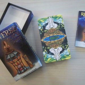 May include: A box of Reverse Scale Tarot cards with a blue and gold design featuring a lantern. The box includes 78 tarot cards and a guidebook. The guidebook is a separate blue and gold booklet with the text "Reverse Scale Tarot" and "The Guidebook".