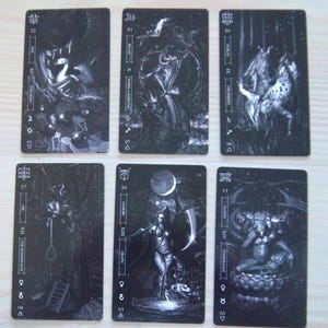 May include: Six tarot cards with dark, gothic illustrations. The cards feature images of Strength, Wheel of Fortune, The Hermit, The Hanged Man, Death, and Temperance. Each card has a unique design with black and white artwork.