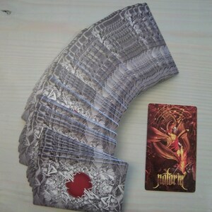 May include: A deck of playing cards with a black and white design featuring a red circle on the back. The cards are fanned out in a semi-circle. A single card is shown to the right of the deck, featuring a red and gold design with the word "notoria" printed on it.