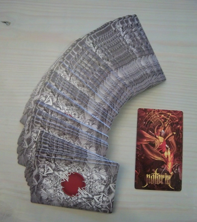 May include: A deck of playing cards with a black and white design featuring a red circle on the back. The cards are fanned out in a semi-circle. A single card is shown to the right of the deck, featuring a red and gold design with the word "notoria" printed on it.
