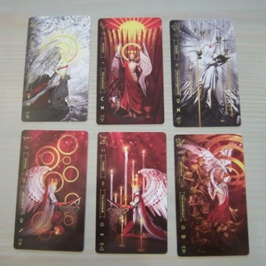 May include: Set of ten tarot cards featuring illustrations of angels with wings and gold accents. The cards are numbered 1-10 and have the names of the angels written on them. The cards are on a gray surface.