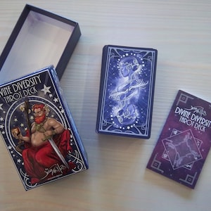 May include: A blue box with the text "Divine Diversity Tarot Deck" and an illustration of a person holding a sword and a cup. The box is open and the tarot cards are visible. The cards have a blue and purple background with stars and an illustration of two people.
