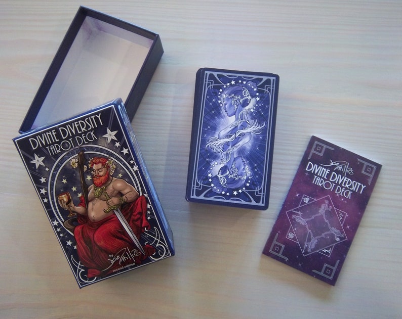 May include: A blue box with the text "Divine Diversity Tarot Deck" and an illustration of a person holding a sword and a cup. The box is open and the tarot cards are visible. The cards have a blue and purple background with stars and an illustration of two people.