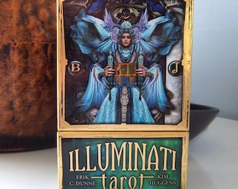 May include: A tarot card box set with a detailed illustration of a figure in blue robes with wings, holding a golden chest. The box has a gold border and the word "ILLUMINATI" is printed on the bottom, along with the word "tarot".