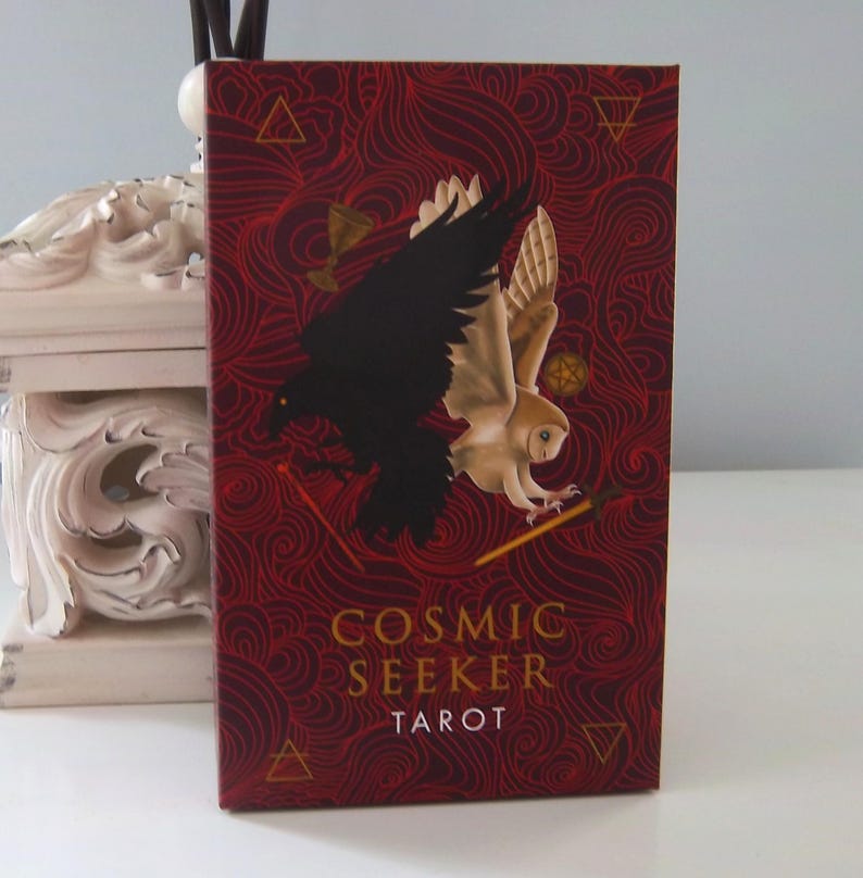 May include: A red tarot deck box featuring a black raven and a white owl in flight against a red background with swirling patterns. The box is labelled in gold lettering with the words "Cosmic Seeker Tarot".