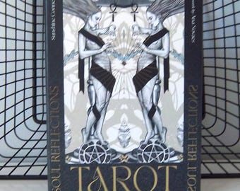 Puede incluir: Una baraja de tarot titulada "Soul Reflections Tarot" con un diseño negro y dorado. La portada presenta dos figuras con cabello suelto, fajas negras y formas geométricas. La palabra "Tarot" está en letras doradas.