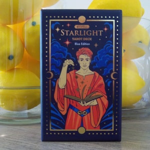 Può includere: Una scatola blu con accenti dorati che presenta un mazzo di tarocchi chiamato "Starlight Tarot Deck - Blue Edition". La scatola ha un design con una persona in una veste rossa che tiene un libro con stelle e una mezzaluna sullo sfondo.