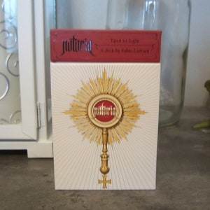 May include: A white box with a red top and gold accents. The box features a gold sunburst design with a red circle in the center that says "natuRla" in gold lettering. The text on the red top reads "Tarot in Light A deck by Fabio Listrani".