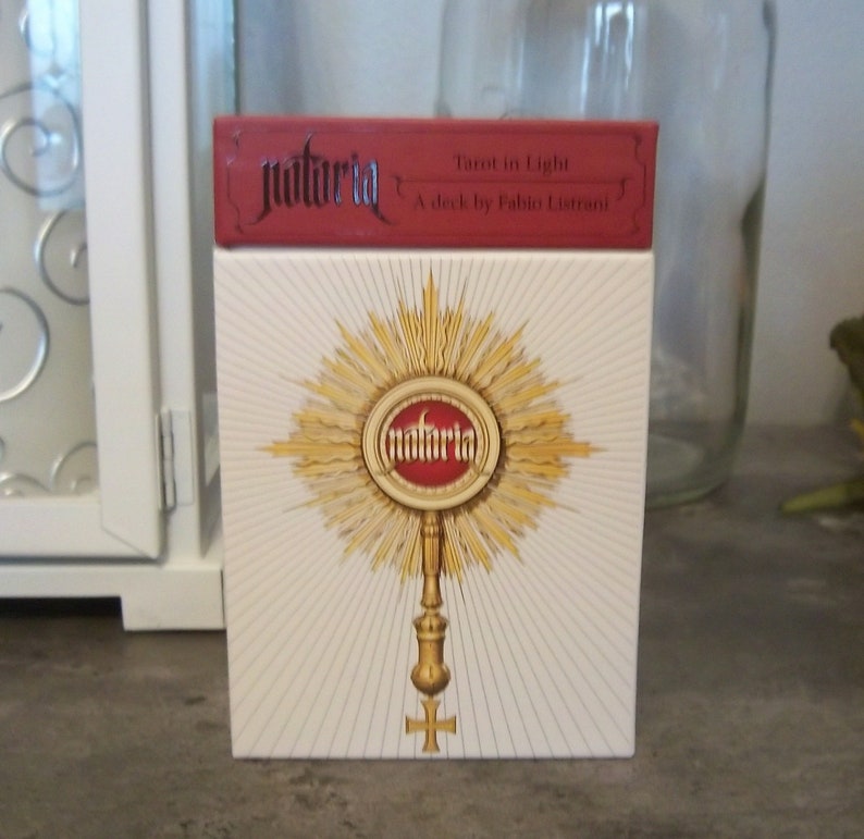 May include: A white box with a red top and gold accents. The box features a gold sunburst design with a red circle in the center that says "natuRla" in gold lettering. The text on the red top reads "Tarot in Light A deck by Fabio Listrani".