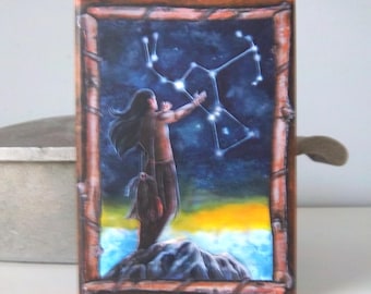 May include: A rectangular wooden box featuring a painting of a person gesturing towards a constellation against a night sky. The image is framed in brown wood. The person stands on a rock formation, with a yellow and blue backdrop.