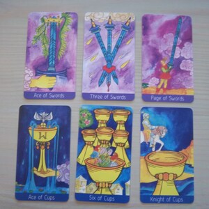 May include: Tarot card spread with six cards, including Ace of Swords, Three of Swords, Page of Swords, Ace of Cups, Six of Cups, and Knight of Cups. The cards are illustrated with watercolor paintings on a purple background.