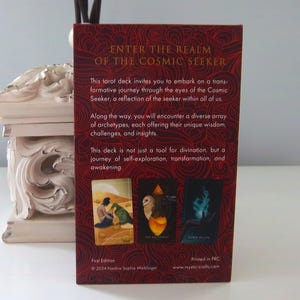 May include: The back of a tarot deck box with the title "Enter the Realm of the Cosmic Seeker". The box is red with gold lettering and features three cards from the deck: "The Sacred Earth", "The Alchemist", and "Return of Life". The text on the back of the box reads: "This tarot deck invites you to embark on a transformative journey through the eyes of the Cosmic Seeker, a reflection of the seeker within all of us. Along the way, you will encounter a diverse array of archetypes, each offering their unique wisdom, challenges, and insights. This deck is not just a tool for divination, but a journey of self-exploration, transformation, and awakening."