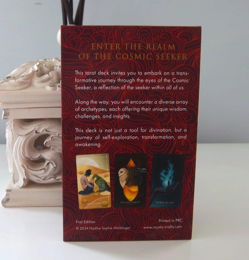 May include: The back of a tarot deck box with the title "Enter the Realm of the Cosmic Seeker". The box is red with gold lettering and features three cards from the deck: "The Sacred Earth", "The Alchemist", and "Return of Life". The text on the back of the box reads: "This tarot deck invites you to embark on a transformative journey through the eyes of the Cosmic Seeker, a reflection of the seeker within all of us. Along the way, you will encounter a diverse array of archetypes, each offering their unique wisdom, challenges, and insights. This deck is not just a tool for divination, but a journey of self-exploration, transformation, and awakening."