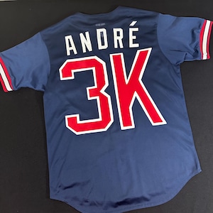 Op de afbeelding: Een marineblauw baseballshirt met rode en witte letters. Het shirt heeft de naam "ANDRE" en het nummer "3K" op de rug.