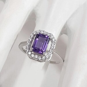 Peut inclure: Une bague en argent ornée d'une pierre précieuse rectangulaire violet foncé, entourée d'un halo de petites pierres claires. La bague est présentée sur une surface blanche, mettant en valeur le design et l'éclat du bijou.