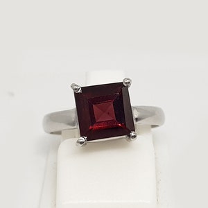 Könnte beinhalten: Ein silberner Ring mit einem quadratischen, dunkelroten Edelstein.