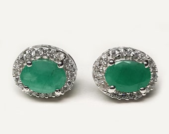 May include: A pair of oval-shaped emerald stud earrings. Each earring features a vibrant green gemstone surrounded by a halo of small, clear, round-cut stones set in a silver-colored metal. The earrings are displayed on a white background.