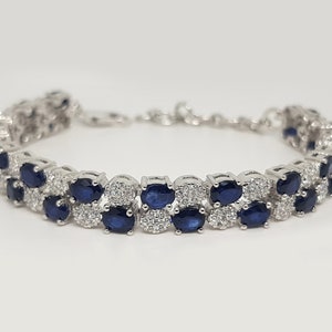 May include: A silver bracelet featuring two rows of oval, dark blue gemstones, interspersed with smaller, clear stones. The bracelet has a delicate chain closure and is set against a white background.