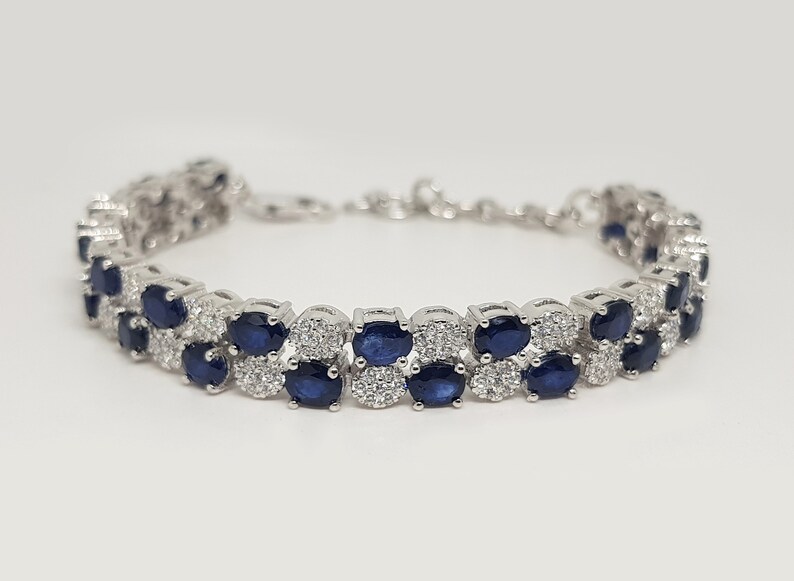 May include: A silver bracelet featuring two rows of oval, dark blue gemstones, interspersed with smaller, clear stones. The bracelet has a delicate chain closure and is set against a white background.