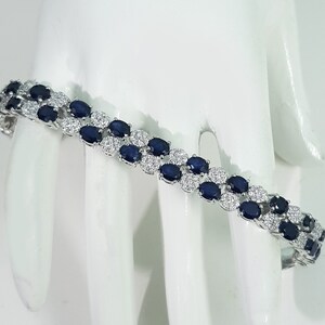 May include: A silver bracelet with two rows of oval, dark blue gemstones alternating with small, clear gemstones. The bracelet is displayed on a white surface, showcasing its elegant design and sparkle.