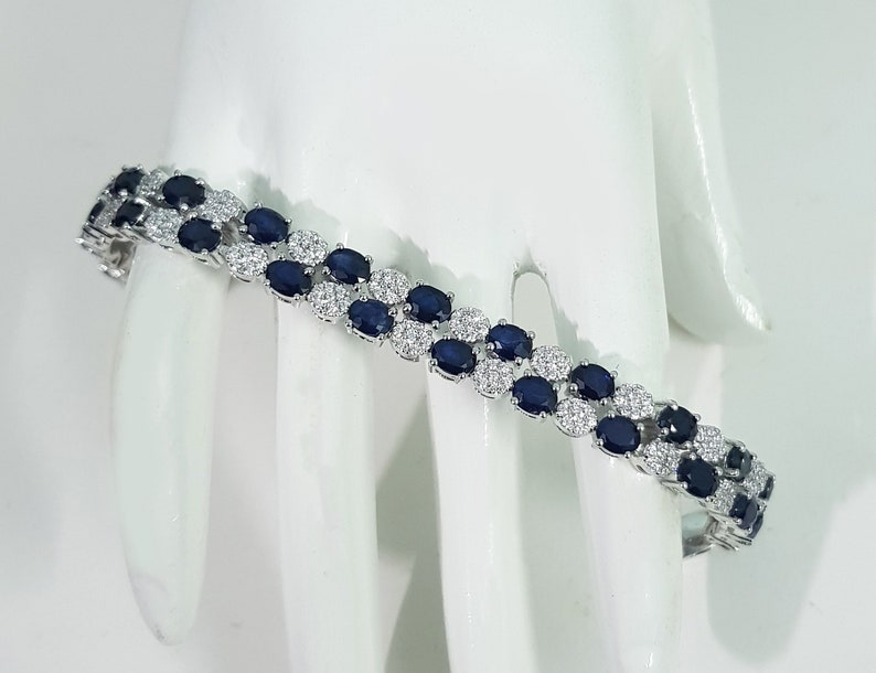 May include: A silver bracelet with two rows of oval, dark blue gemstones alternating with small, clear gemstones. The bracelet is displayed on a white surface, showcasing its elegant design and sparkle.