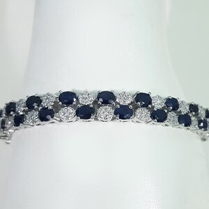 May include: A silver bracelet featuring two rows of oval, dark blue gemstones alternating with clusters of small, clear gemstones. The bracelet is set against a white background, highlighting the sparkle of the gems.
