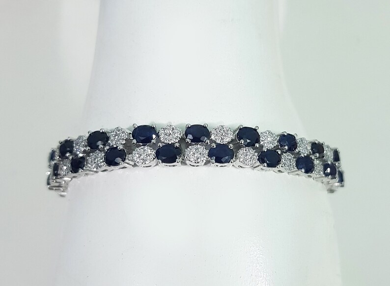 May include: A silver bracelet featuring two rows of oval, dark blue gemstones alternating with clusters of small, clear gemstones. The bracelet is set against a white background, highlighting the sparkle of the gems.
