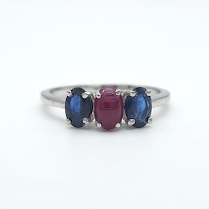 May include: A three-stone ring featuring a central oval cabochon ruby flanked by two oval faceted blue sapphires. The gemstones are set in a polished silver-toned metal band with four-prong settings for each stone, creating a classic horizontal arrangement.