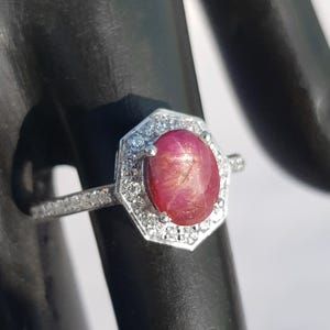 May include: A silver ring with a red star ruby gemstone surrounded by a halo of smaller clear gemstones.