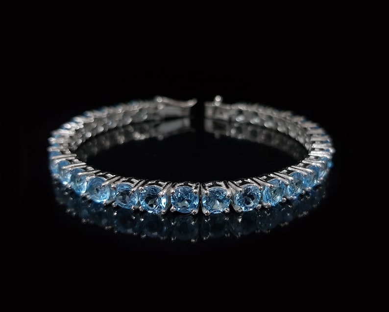 May include: A silver bracelet with a row of light blue round gemstones. The bracelet is on a black background.